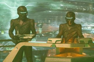 two humanoid aliens wearing black armor and helmets hold laser rifles