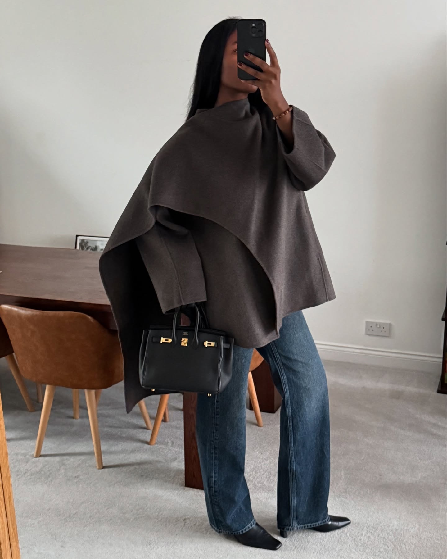 British style influencer Marilyn Nwawulor Kazemaks poses for a mirror selfie wearing a brown scarf coat, Hermes bag, bootcut jeans, and black kitten-heel boots