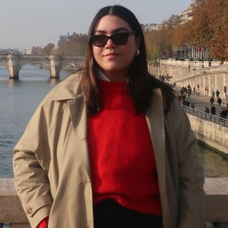 Woman wearing red sweater, classic khaki trench coat, black sunglasses, standing on Paris bridge.