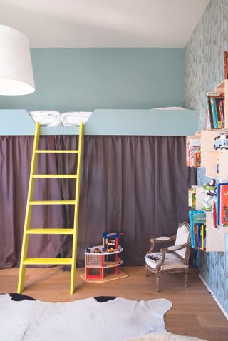 boys bedroom with pale blue walls and wallpaper, brown curtain underneath high bed, yellow ladder, bookcase