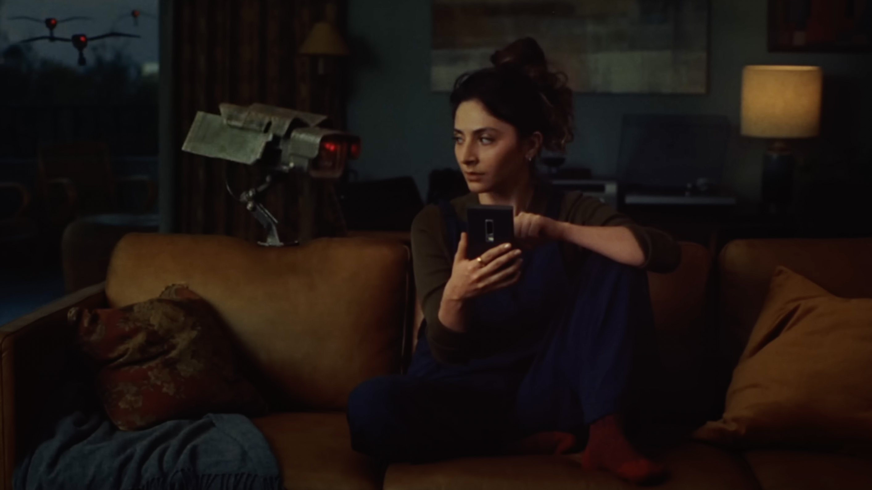 A woman sits on a couch and scrolls through her phone, with a security camera next to her