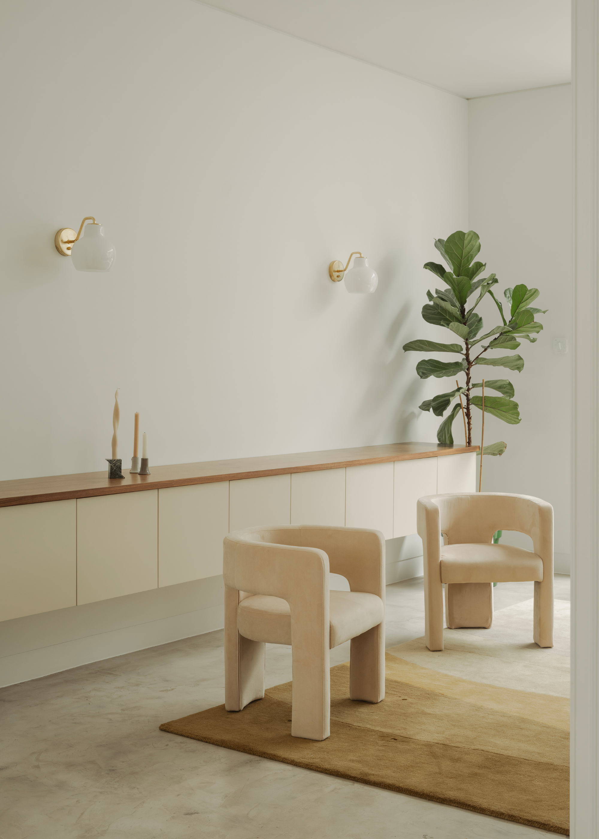A living room with a pair of cream sofa chairs, a fiddle leaf fig tree plant, a pair of wall sconces, a floating sideboard and a trio of candles