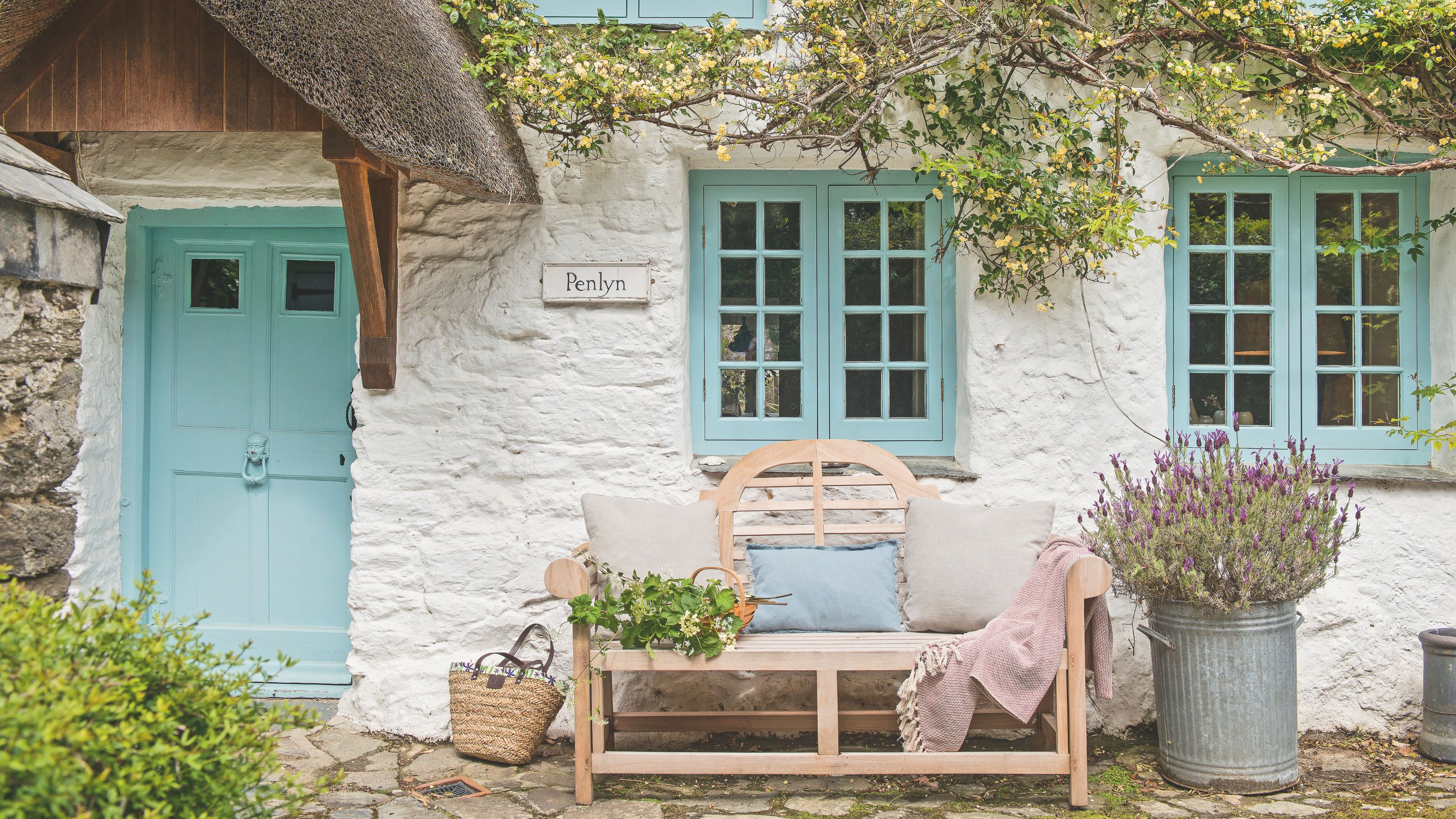White painted cottage frontage with bright blue painted front door and windows. There's a wooden bench with cushions on it in front of the cottage