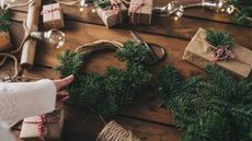 Wooden table showing Christmas wreath being made with fresh spruce on a willow base, with presents wrapped in brown paper