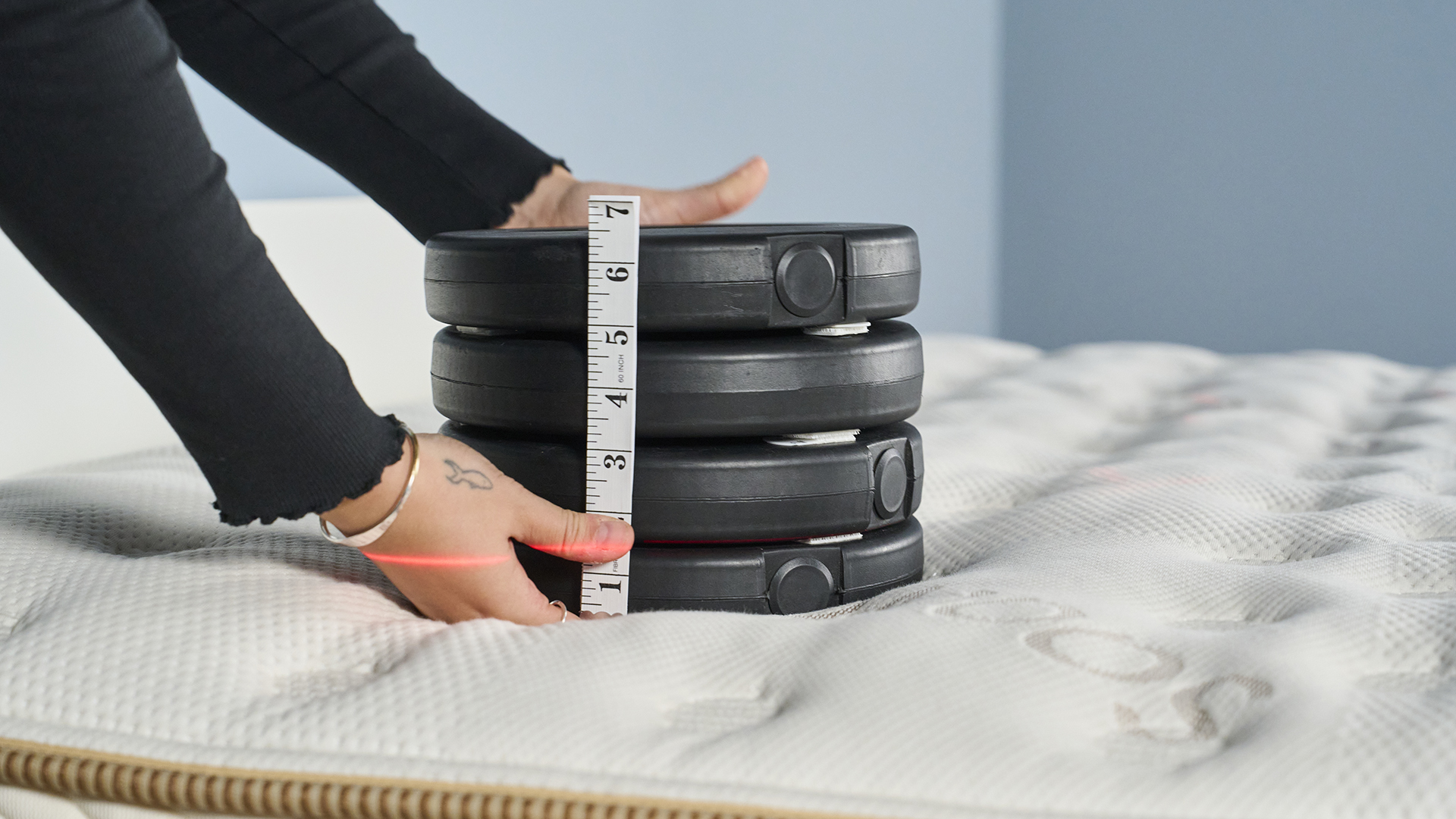 Pressure relief lab test image shows a stack of black weights being placed on the Saatva Classic mattress, in the left hand shoulder area, to measure material indentation and recovery. A read laser line points to the original starting point of the weights so we have a visual record of how much the weight is sinking down, in addition to our two measuring tapes used to record indentation.