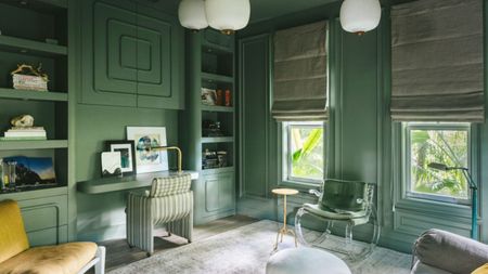 A jade green home office with the desk up against the wall with a paneled design on the cupboard doors above it, two windows with roman blinds, and multiple seats and poufs