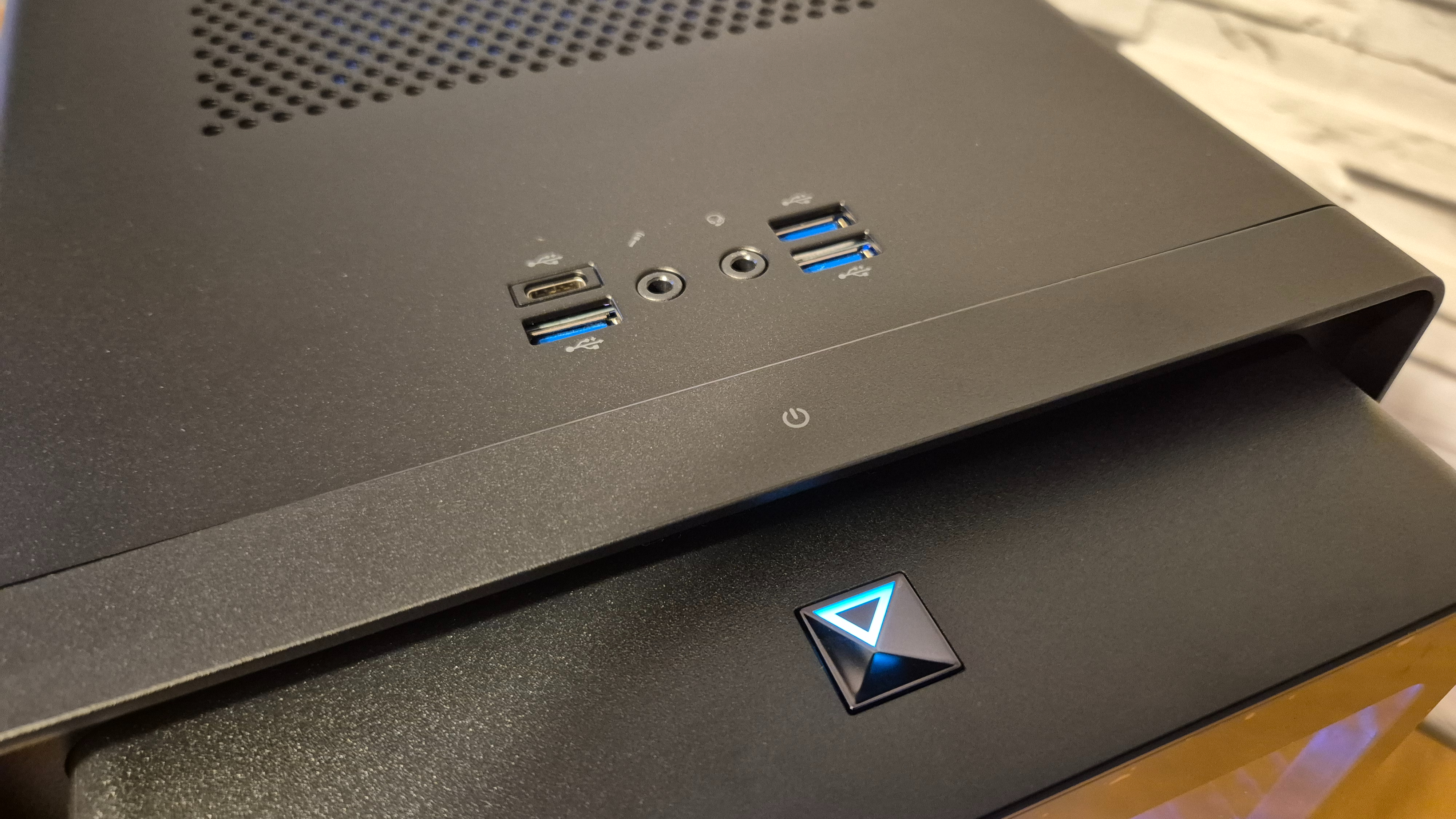 A close up of the buttons and ports on the front of an Acer Predator Orion 5000 gaming PC as it sits on top of a wooden surface infront of a white brick effect wall