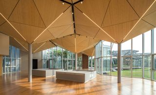 The interior section of the building featuring geometric shaped ceiling made from bamboo and held up with grey steel pillar. Glass wall panels with a view of the garden