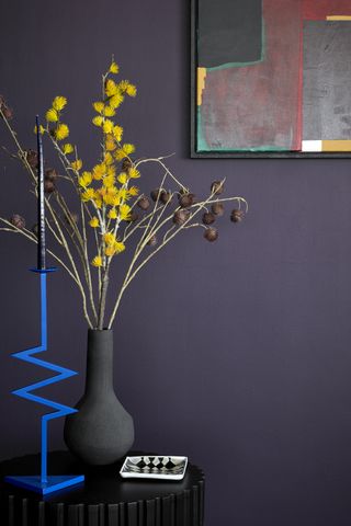 A small portion of a room painted purple, complemented by a grey vase and an electric blue candle stand