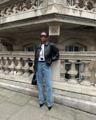 London woman wears baggy light wash jeans with a black leather bomber jacket, skinny belt, and patent leather heels.