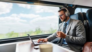 A man in a suit sitting on a train that appears to be moving. He&#039;s using a laptop and headphones