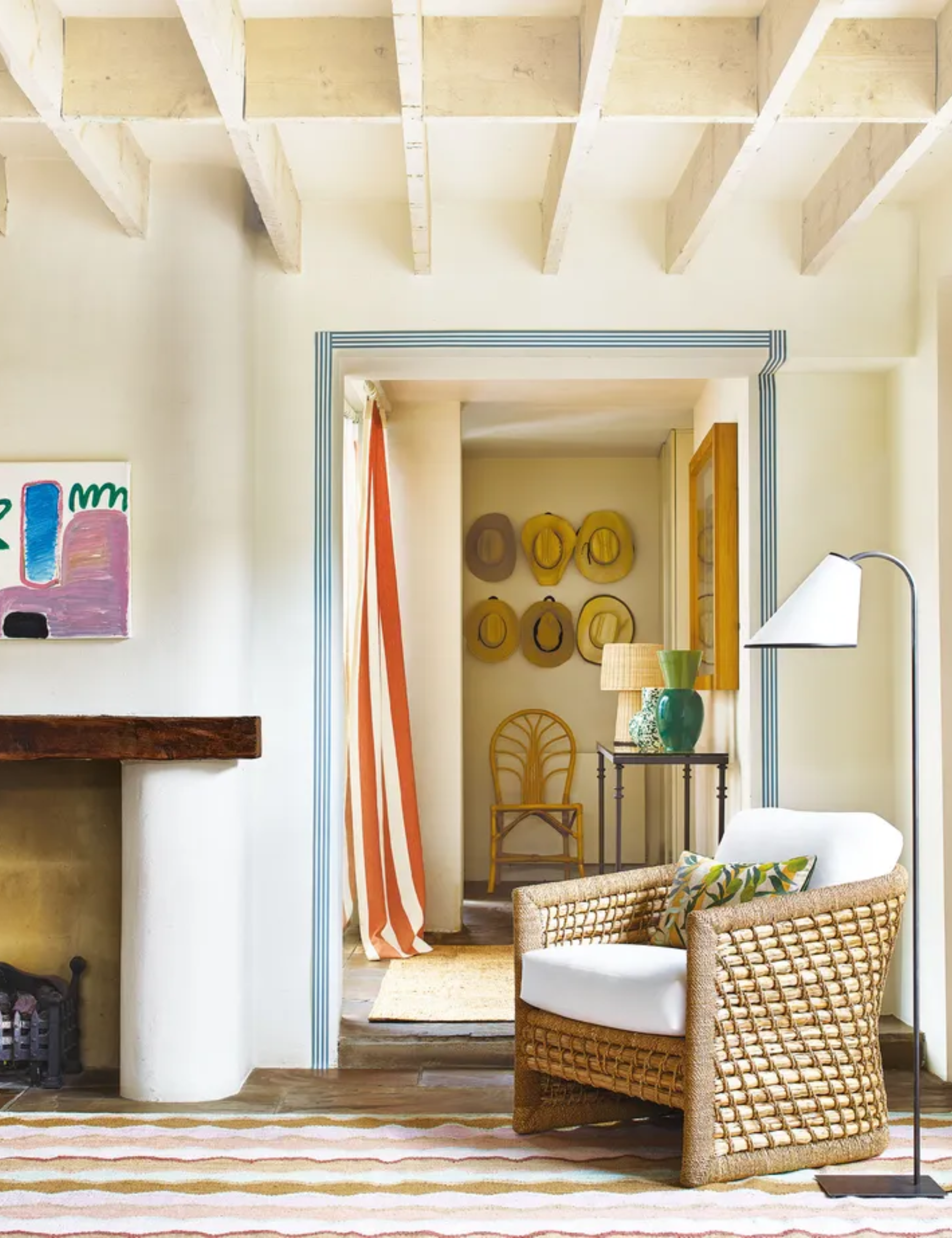 Modern, country cottage with coffered wooden beams, cream painted walls, armchair with floor lamp beside, patterned rug, fireplace with modern artwork above, looking through to the hallway with side table, decorative hat display on walls, wooden chair