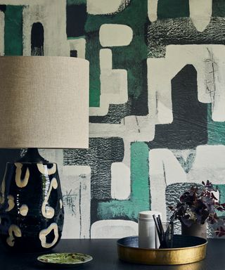 Abstract black, green and cream wallpaper behind a black side table and ceramic lamp.