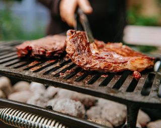 Two steaks being cooked on outdoor grill BBQ with one being upturned by person with a cooking utensil