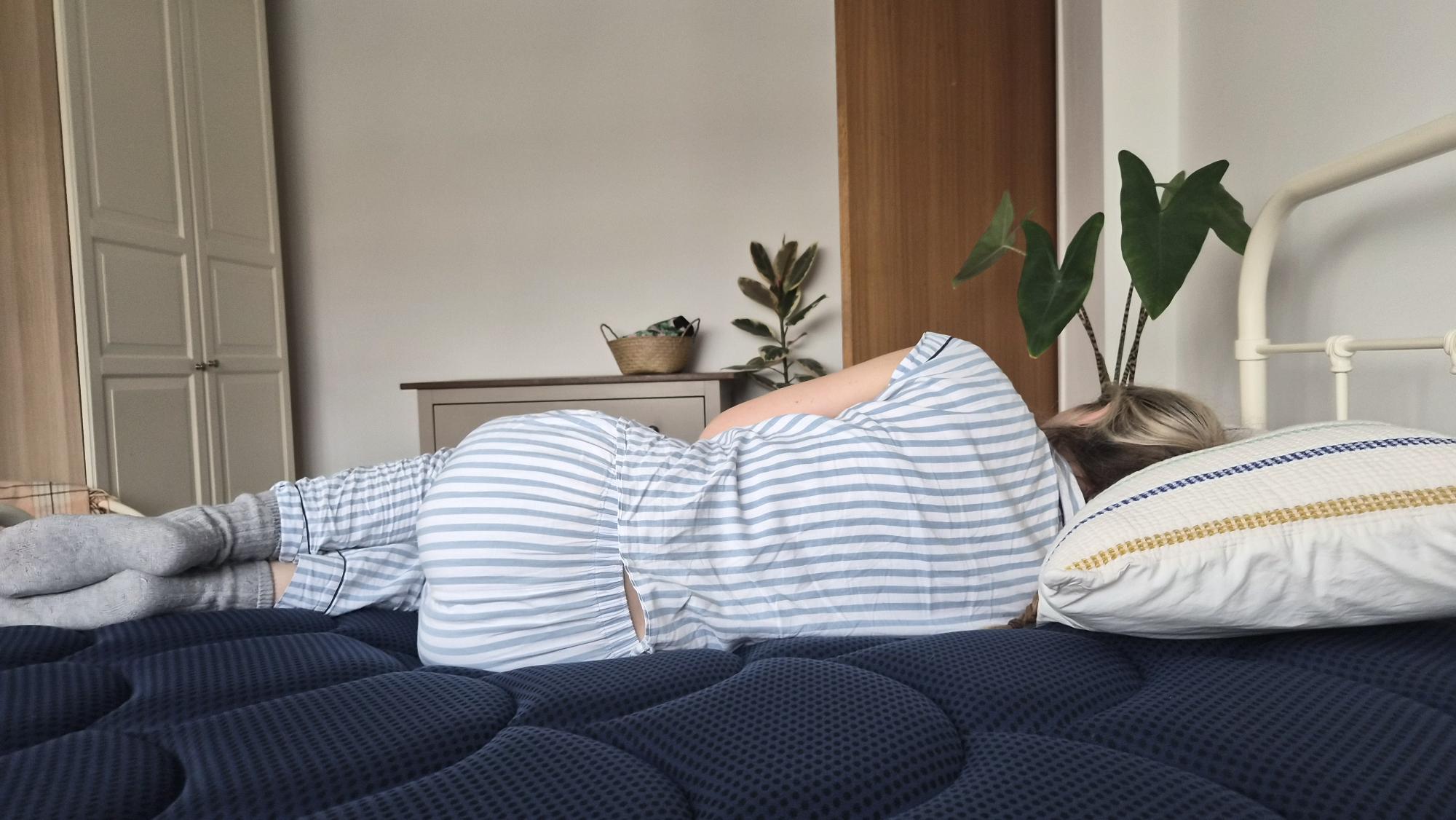 The image shows a light haired mattress tester wearing blue and white stripey pyjamas lying on her side on a dark blue mattress with her back to the camera. 