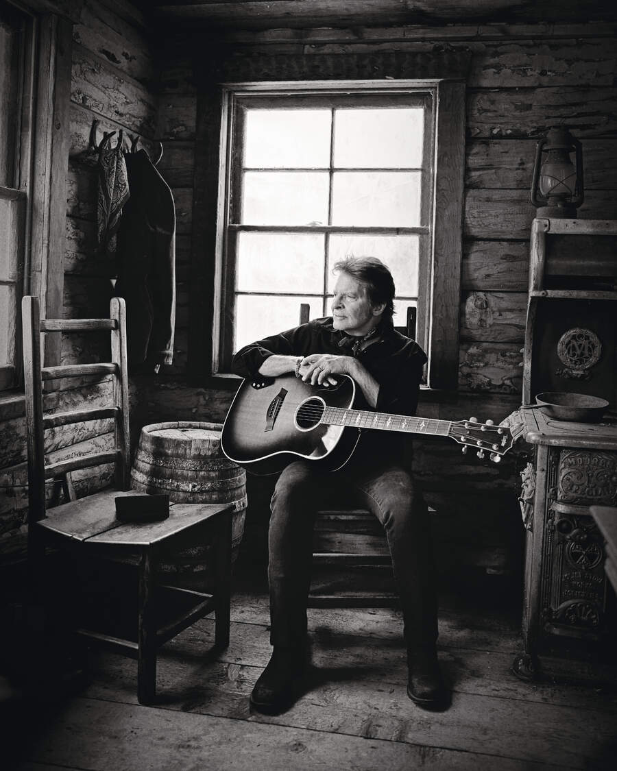John Fogerty sitting in a log cabin with an acoustic guitar