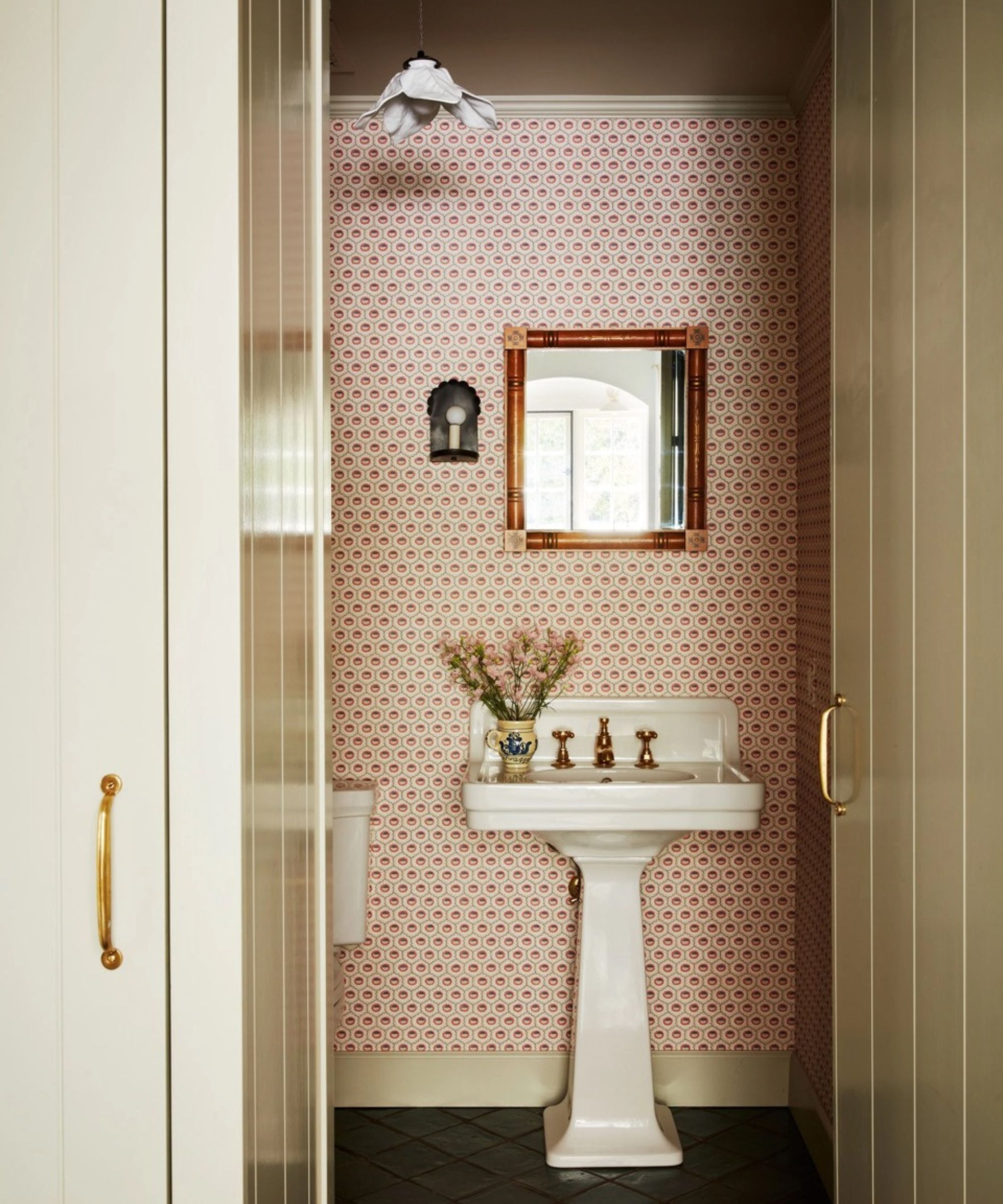 A cozy powder room viewed through a doorway, featuring pink and white geometric wallpaper, a classic white pedestal sink with brass hardware, and a small wood-framed mirror. A floral pendant light and a wall sconce provide warm, soft lighting.