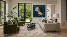 A modern living space with a green and beige sofa facing each other with a black two-layered thin coffee table in the center styled with a vase of foliage, books and candles; and there is a large artwork of the side of a girl's face on the wall next to black-framed doors leading to the garden