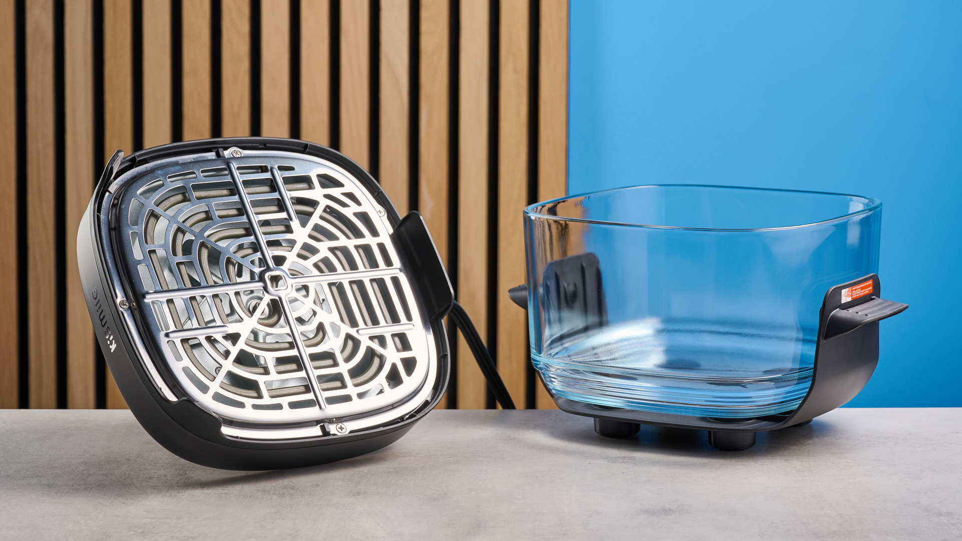 the kismile roasti air fryer with a glass cooking container photographed against a blue and wood background, showing the heating element lid