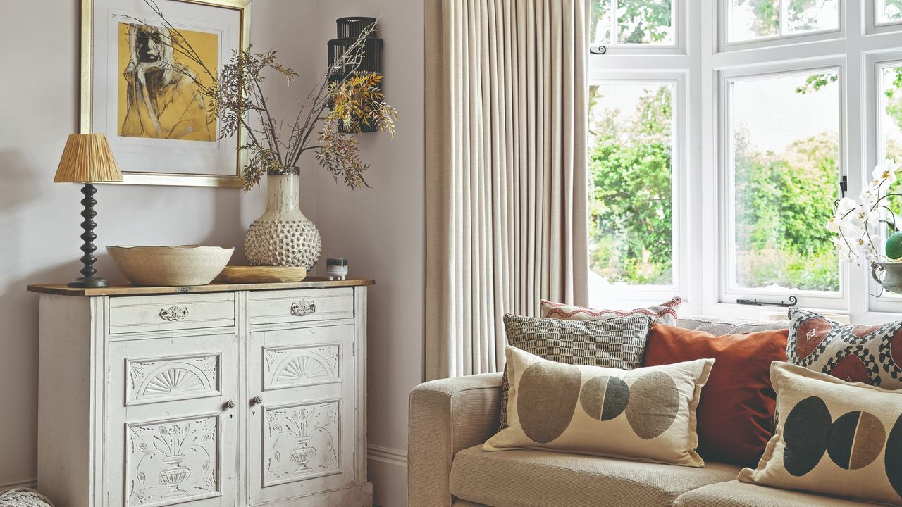 A light, bright living room with a beige sofa with mismatched cushions and a table lamp on a sideboard with a wall light to the side of it