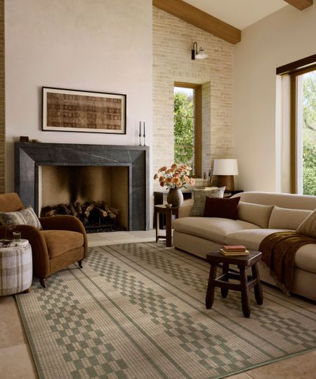 Warm neutral living room with beamed ceiling, a black marble fireplace, a beige curved sofa, a chequered green and neutral rug, a brown velvet armchair, and a rustic wooden stool.