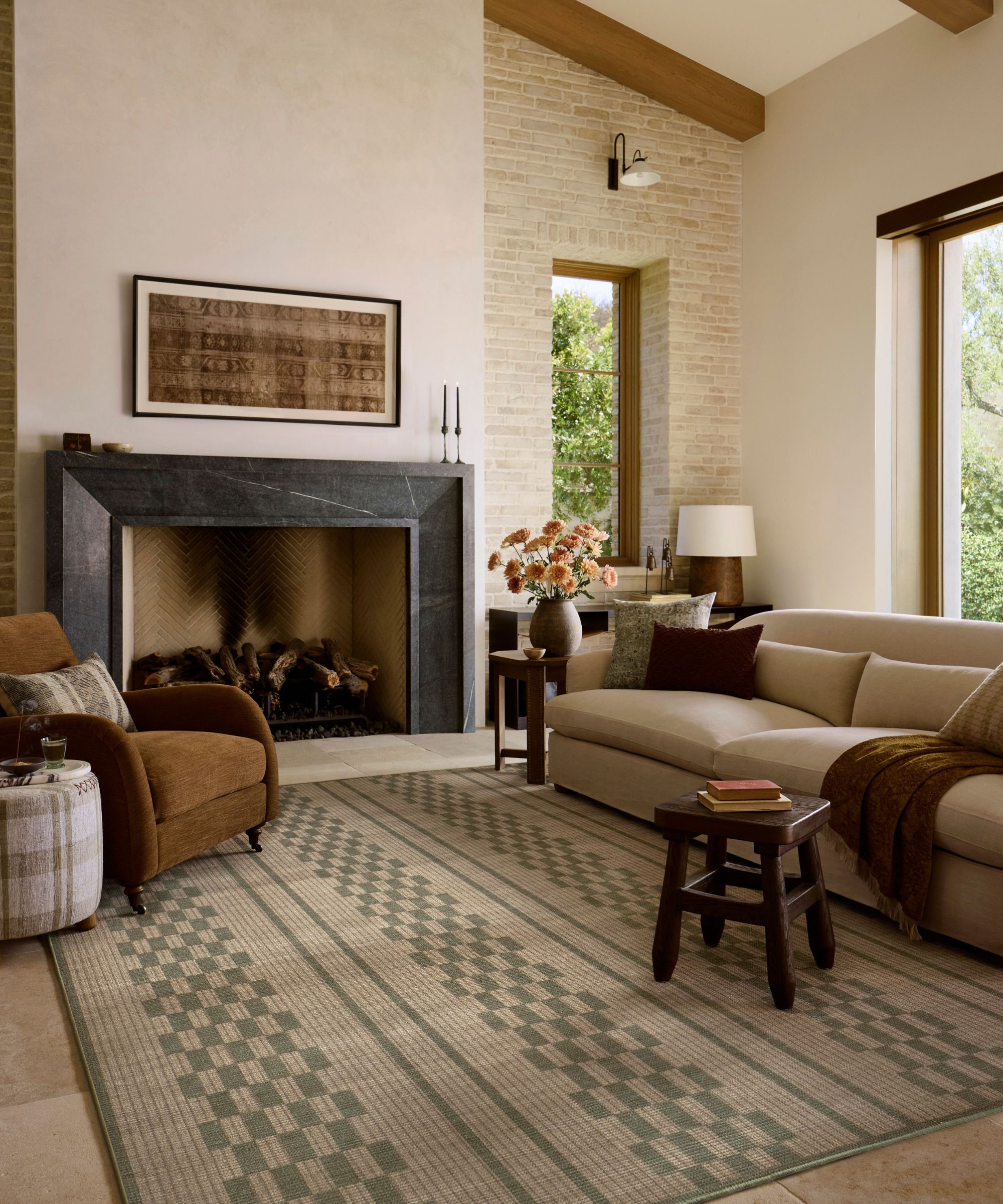Warm neutral living room with beamed ceiling, a black marble fireplace, a beige curved sofa, a chequered green and neutral rug, a brown velvet armchair, and a rustic wooden stool.