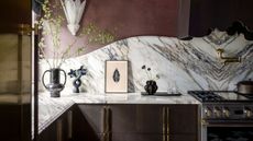 a dark red maximalist kitchen with a curved marble backsplash, that bookmatches in the center, under the cooker hood. 