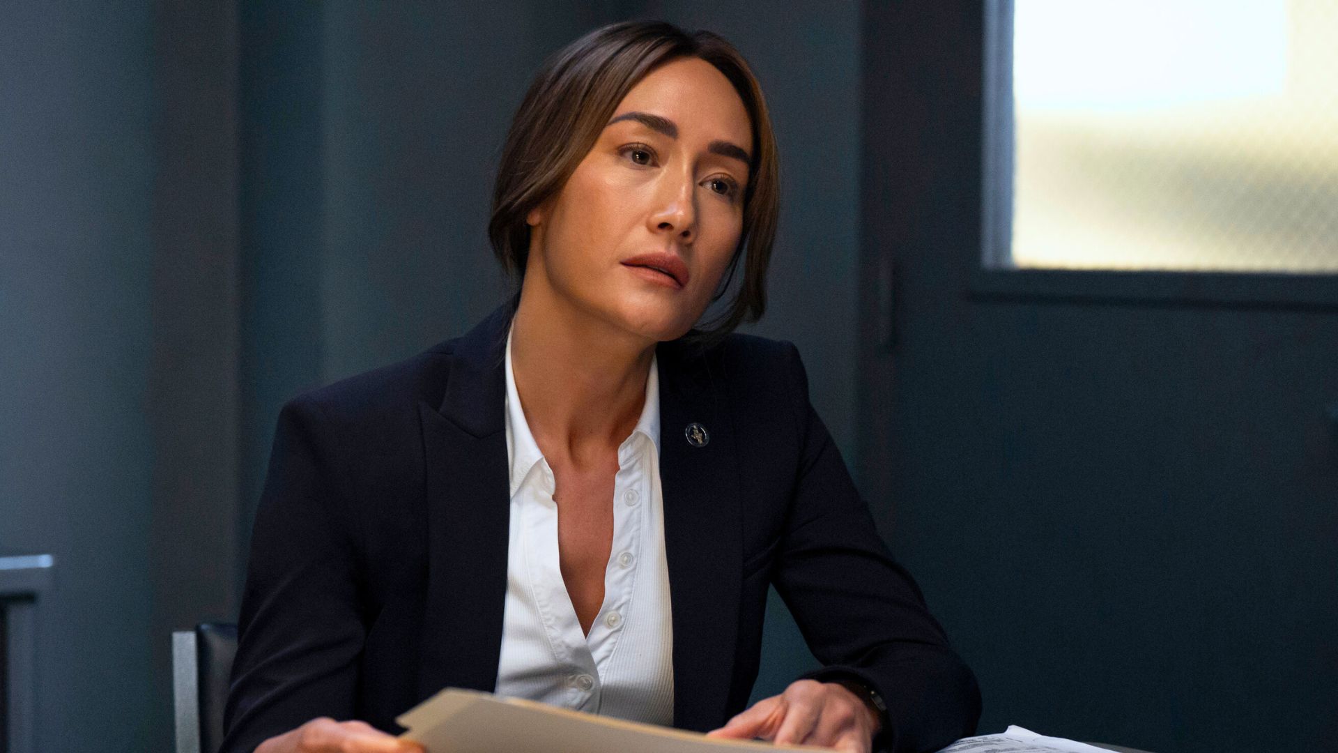 Maggie Q sits at a desk holding a brown folder, looking at something off screen