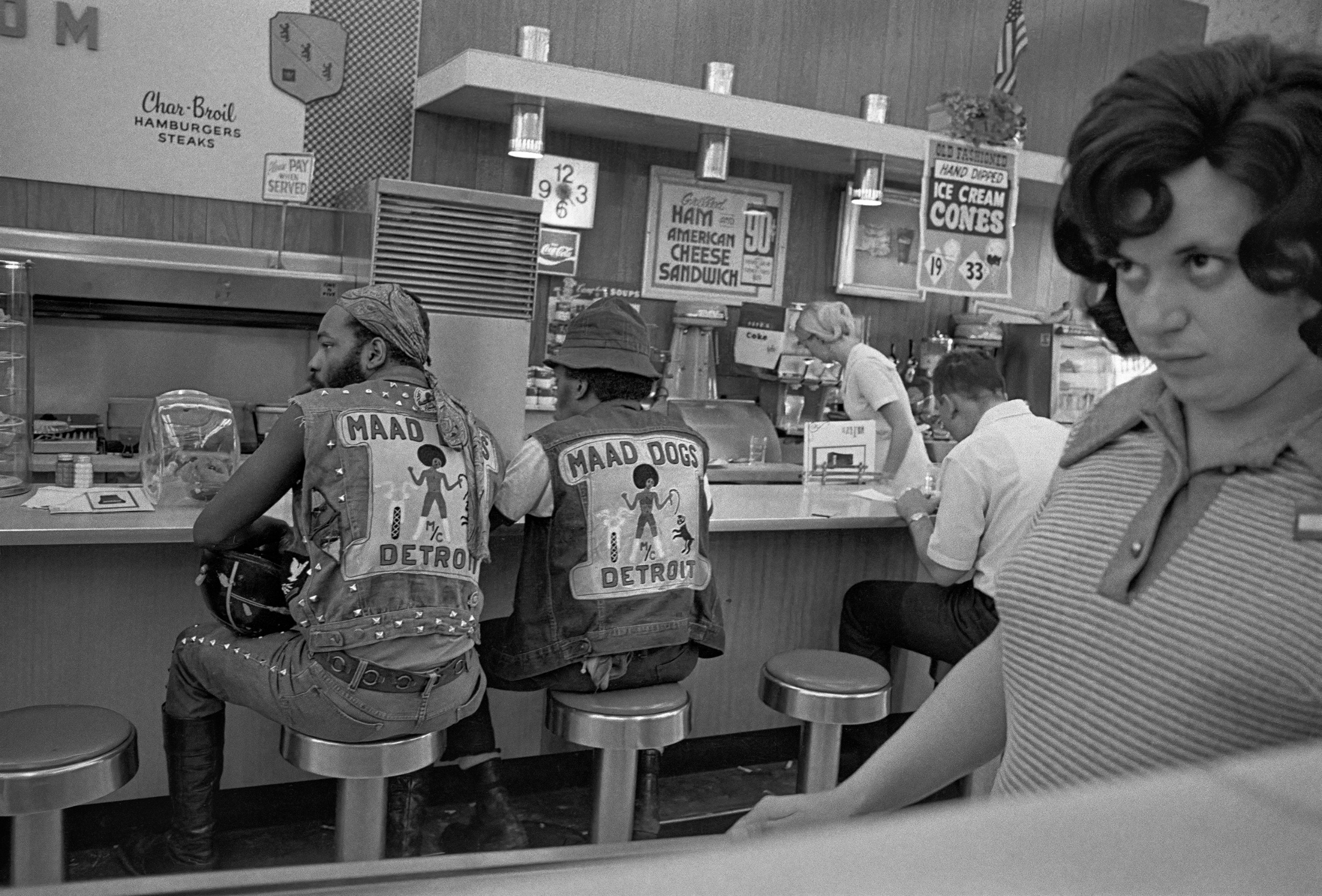 Two men in studded denim vests bearing "Maad Dogs Detroit" motorcycle club patches sit at a diner lunch counter, while a woman in the foreground glances sideways at the camera.