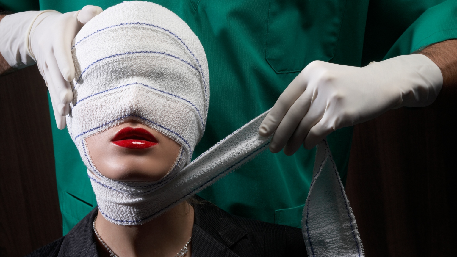 a woman in red lipstick with a head wrap and a surgeon behind her wrapping her face