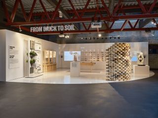 From brick to soil exhibition in tallinn, grey floor, red metal girder ceiling structure, white signage lettering, white curved setting to display items, brick wall, lighting