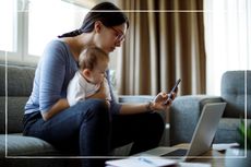 worried mother with baby on her lap looking at phone and laptop while sitting on the sofa at home