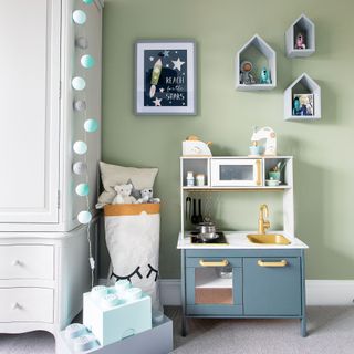 Sage green nursery with blue play kitchen, box shelves on the wall and a white wardrobe