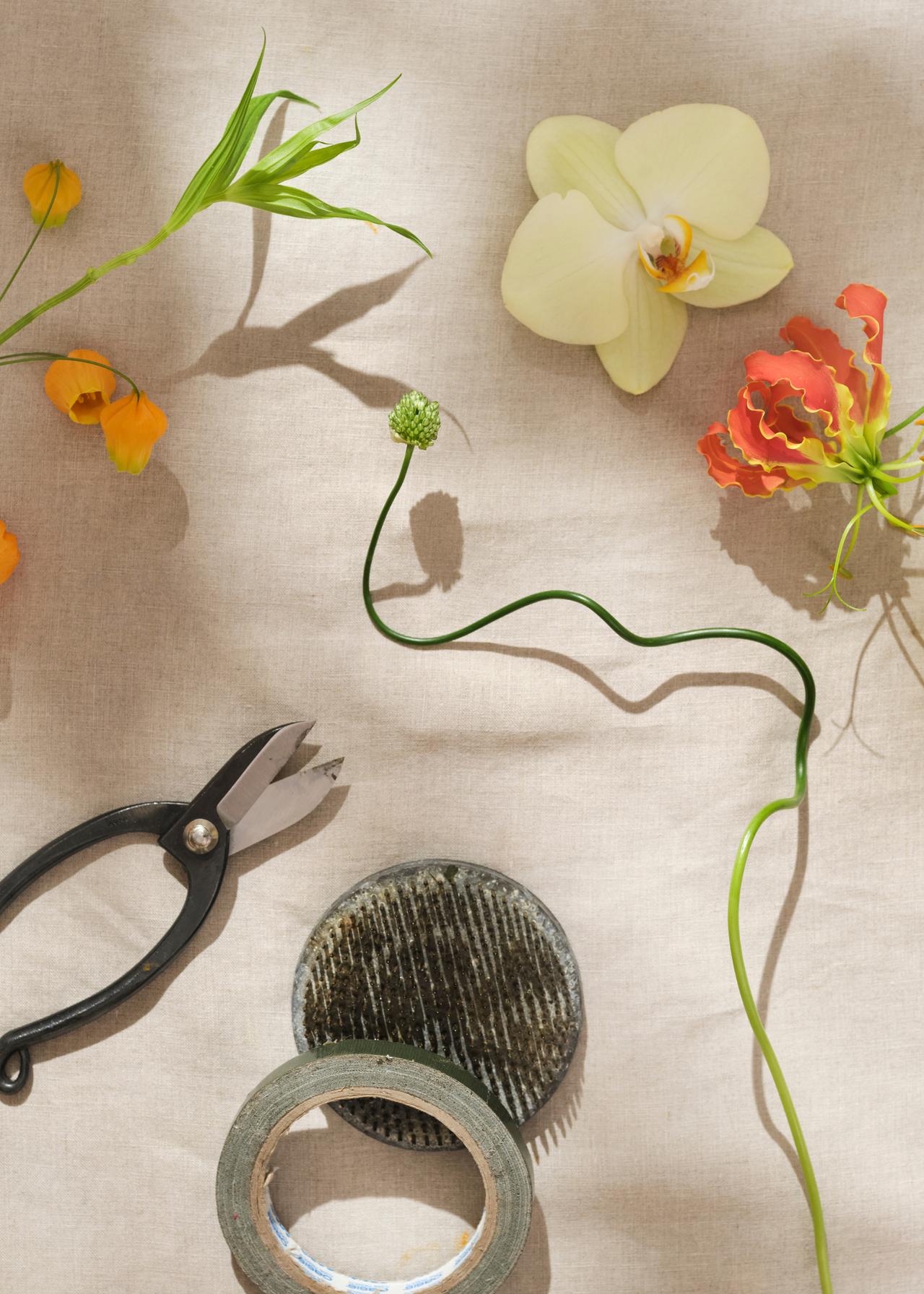 A table with floral secateurs, a kenzan flower frogs, cut flowers and stems, and a roll of tape