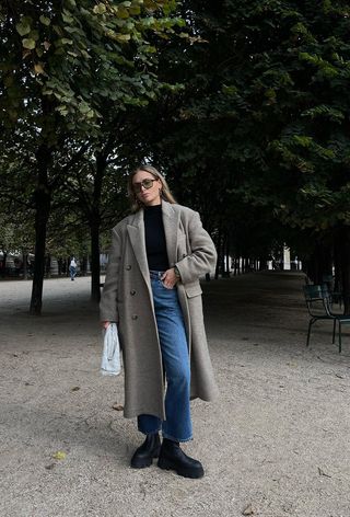 how to wear Chelsea boots is shown in a photo of a woman standing in a park wearing a long tan coat over a black sweater with straight-leg cropped jeans, black chelsea boots, and a white clutch bag