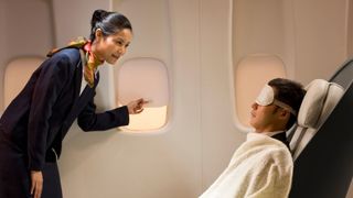 A young flight attendant wearing navy uniform and a colourful necktie shuts a plane window so a male passenger wearing a white eye mask can get some sleep while travelling.