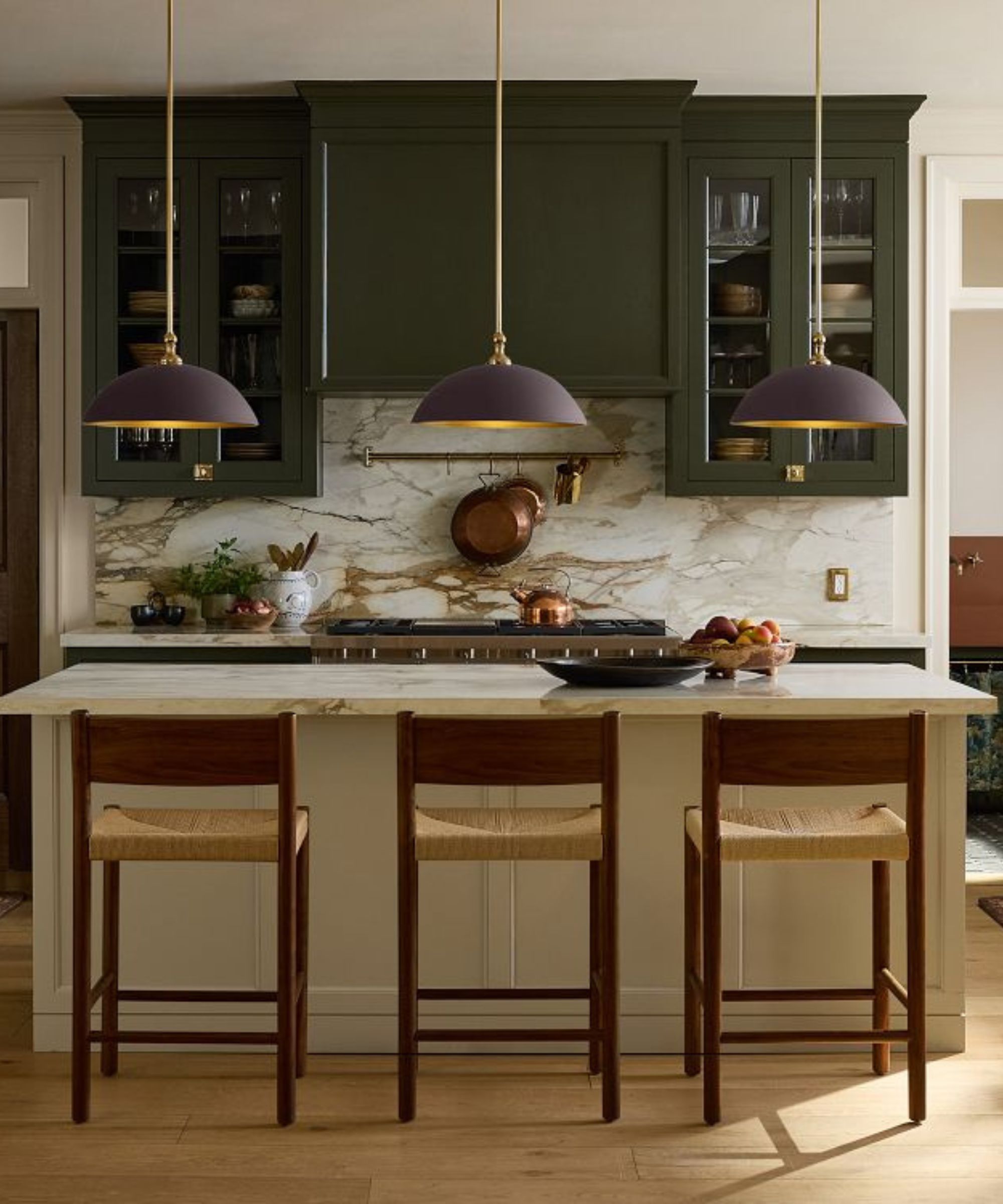 A classic kitchen with green upper cabinets, a cream island, marble countertops, and three brass and mulberry pendant lights