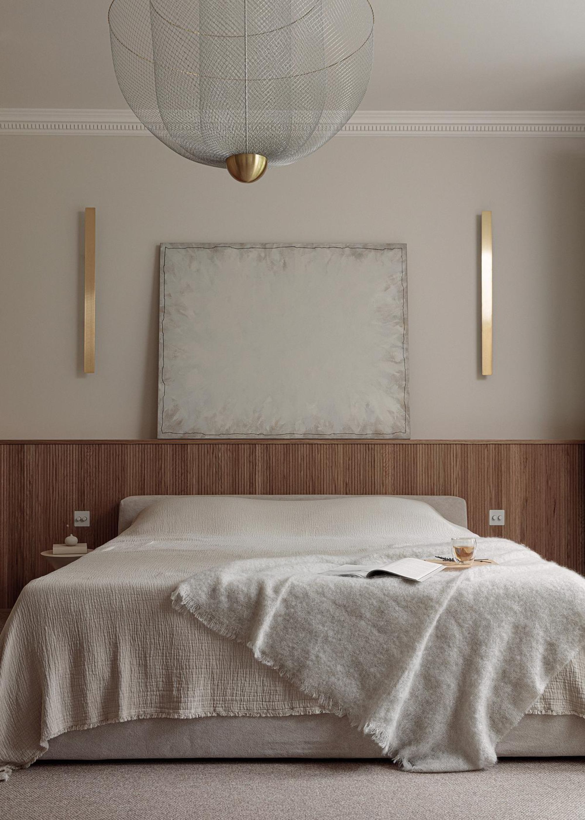 A bedroom with a scandi design, and on top of the bed is an open book and a wooden board, and a glass with a beverage inside