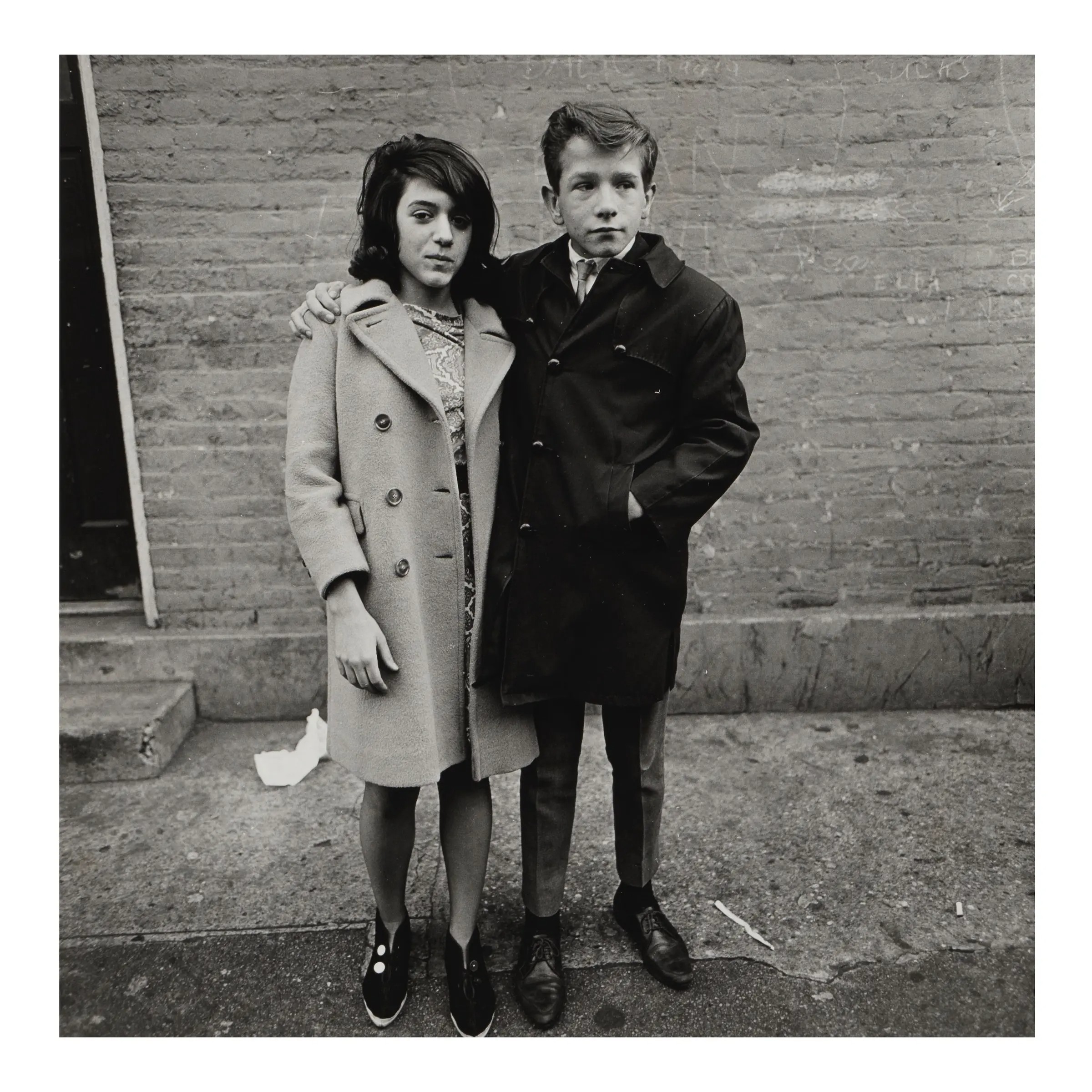 A black and white photograph shows a young girl in a light-colored wool coat and a young boy in a dark trench coat standing close together on a city sidewalk in front of a brick wall.