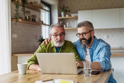 Mature son helping father to manage his finance, teaching how to work with internet baking and shopping online. Handsome son supporting dad, technology and digital literacy.