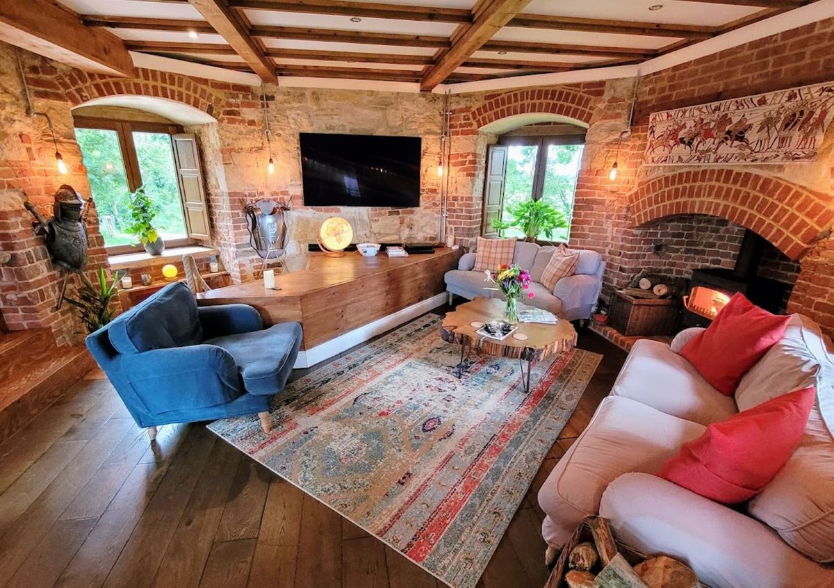 The living room has traditional dark furniture which sit opposite the open fireplace and TV hanging on the wall which is also opposite the staircase. The roof has wooden beams and the fireplace is brick
