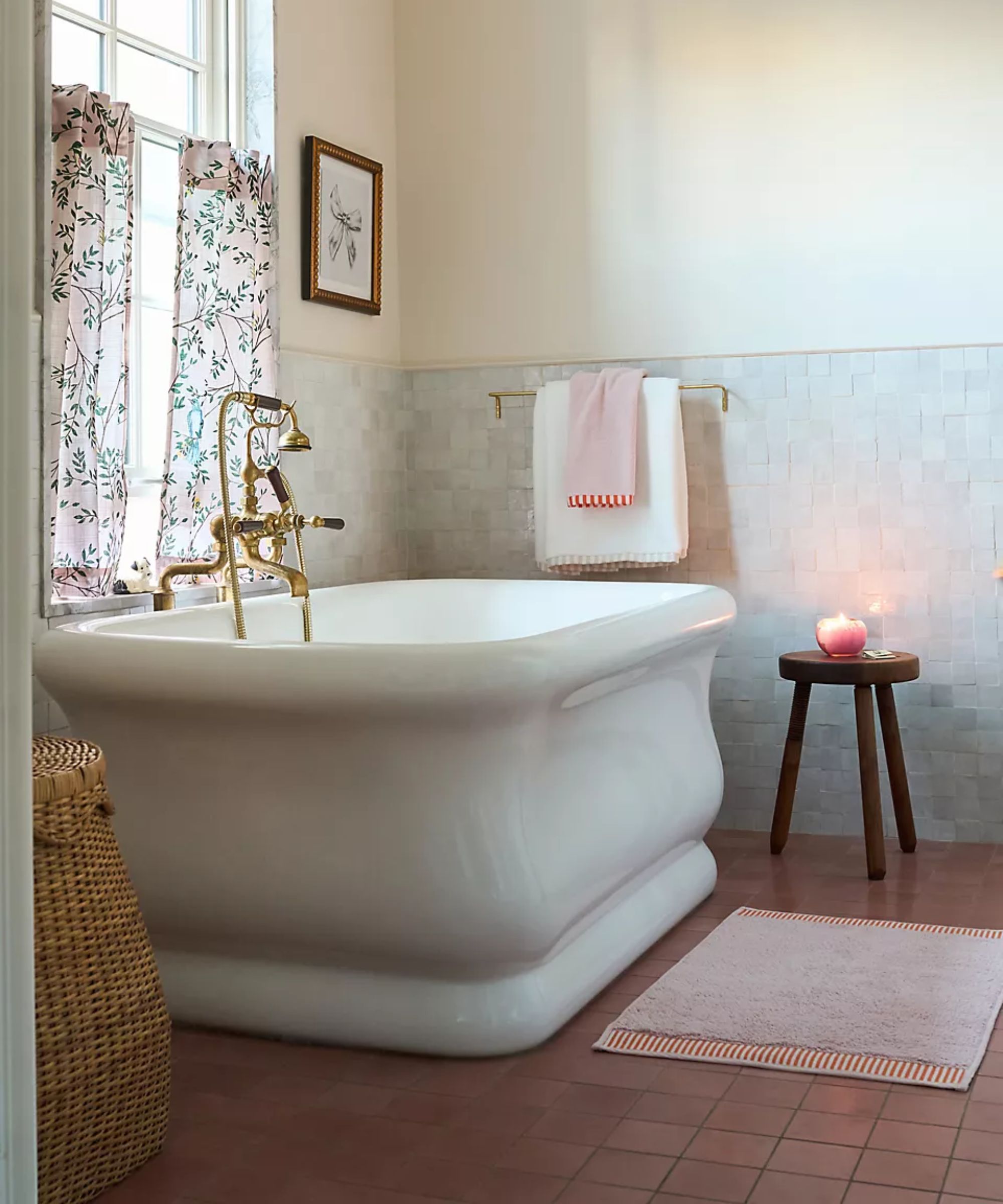 anthropologie styled bathroom with terracotta floor tiles, zellige wall tiles, a large sculptural freestanding bath tub, wooden stool with candle and a floral cafe curtain at the window