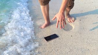 Someone bending to pick up a phone on a beach as a wave washes towards it