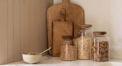 A white kitchen counter with wooden cutting boards leaning against the wall, three glass food jars with wooden lids filled with dried food goods, and a stonewear breakfast bowl beside them