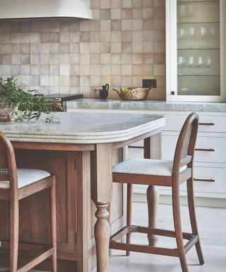 Kitchen with curved wooden island with marble top and beige tiles and white cabinetry with ribbed glass