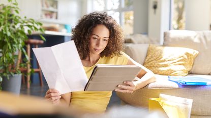 A young woman looks at paperwork while sitting on her living room floor with her laptop.