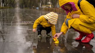 A toddler boy and his mother play in the rain on a paved walkway, both wearing yellow raincoats