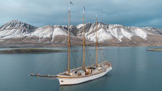 A majestic tall ship anchored in calm waters, with snow-capped mountains towering in the background under a moody sky