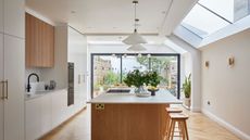 A white and oak kitchen with lighting hanging on top of an island. On the island is an arrangement of green foliage 