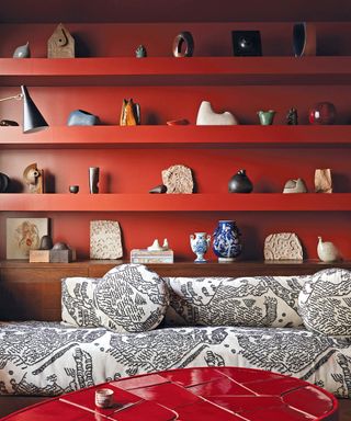Bright red living room with built in shelving, decorated with a large collection of decorative ornaments and objects, black and white patterned sofa in foreground